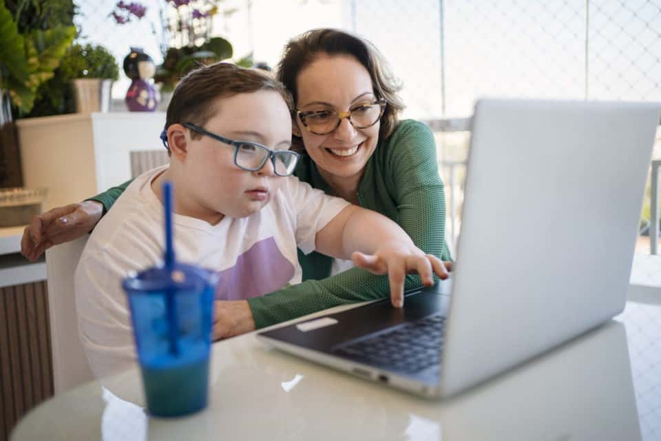 Mother helping son with down syndrome in homeschooling. Distance learning concept.