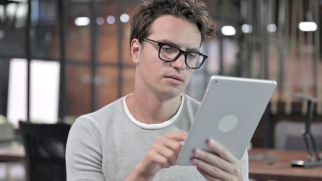 A man with glasses reading something on an iPad. His finger is touching the screen and he is looking at the screen with a neutral expression on his face.