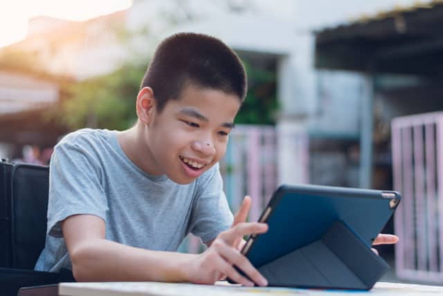 A boy in a wheelchair with an iPad on his tray. He is smiling and has his finger raised ready to touch the iPad.
