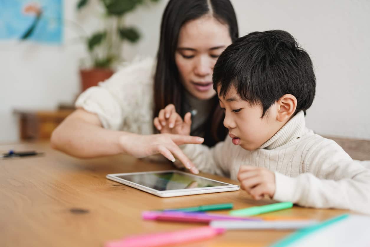An Asian woman sitting with an Asian boy at the table. There are coloured pencils and an iPad on the table. The Asian woman is pointing to the iPad that they are both looking at.