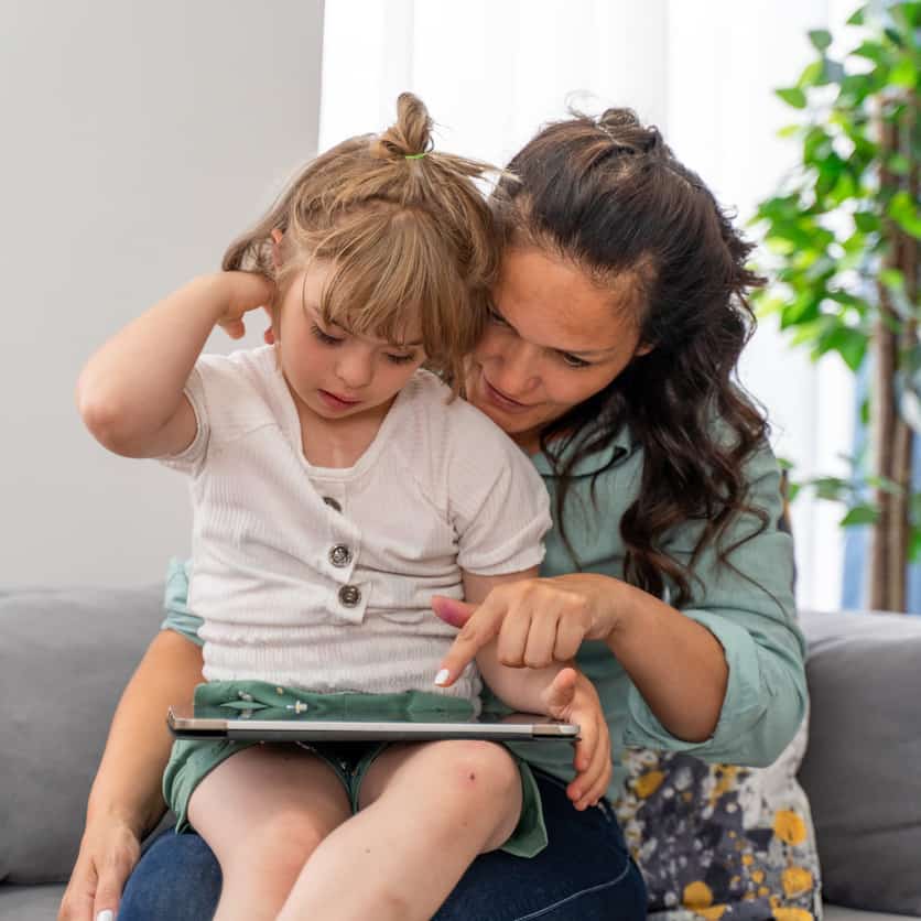 A white young girl sitting on her mum's lap looking at an iPad.
