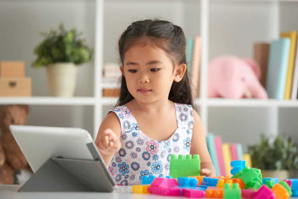 A young asian girl sitting at a table with plastic blocks and an iPad. She is pointing to the iPad.