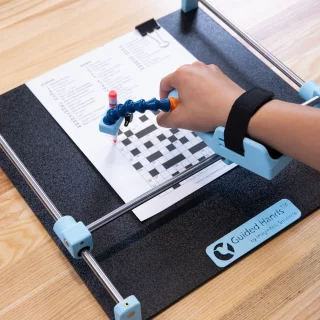A person's hand setup in a Guided Hands assistive device. They are using the Guided Hands to complete a crossword.